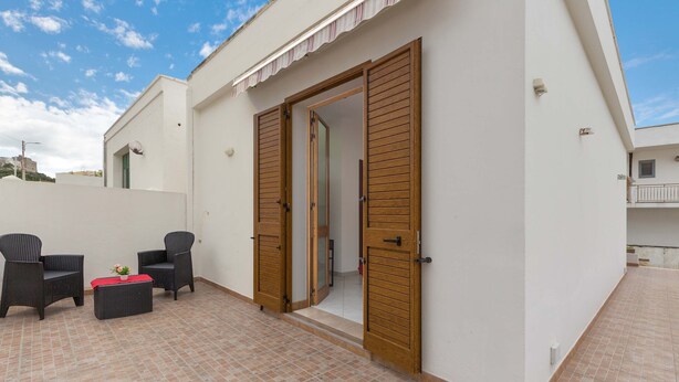 A patio area with two wicker chairs, a small table with a red cushion, and a wooden door leading to an interior space. - 2214 Villetta Enea - Mono 1 by Barbarhouse (Castro)