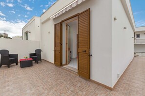 Un'area patio con due poltrone in vimini, un tavolino con un cuscino rosso e una porta in legno che dà su un ambiente interno.