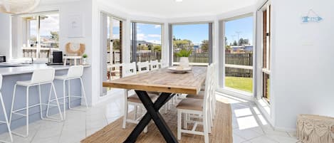 Une cuisine lumineuse avec une table à manger en bois, des chaises blanches et une vue sur le jardin à travers de grandes fenêtres.