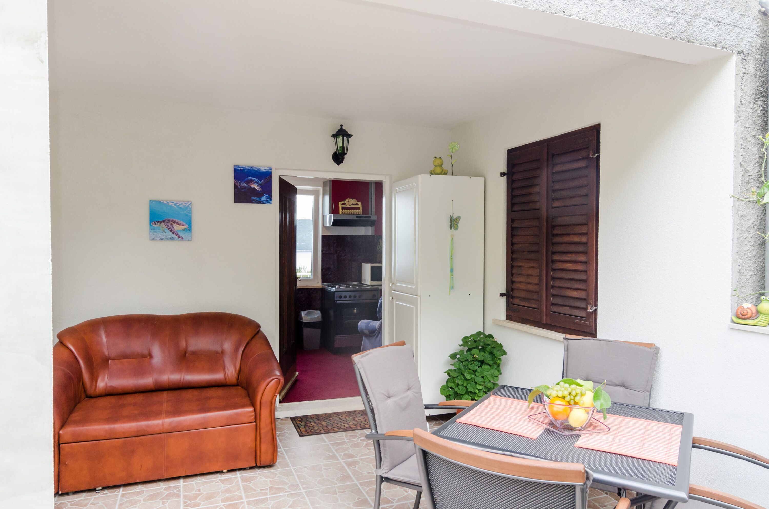 A cozy living area with a brown leather sofa, a dining table with a fruit bowl, and a kitchen in the background.
