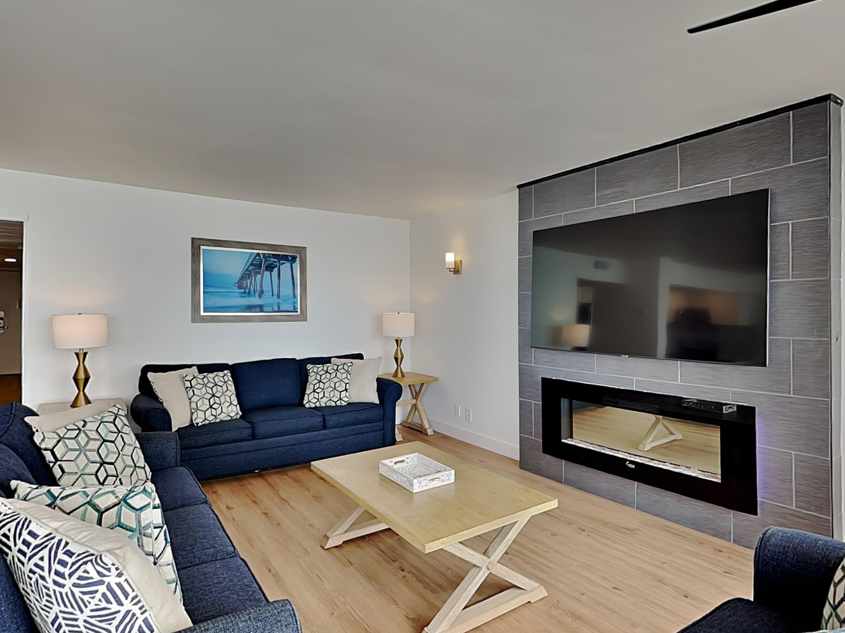 Een moderne woonkamer met een blauwe bank, een salontafel, een flatscreen-televisie aan een grijze muur en een open haard eronder.