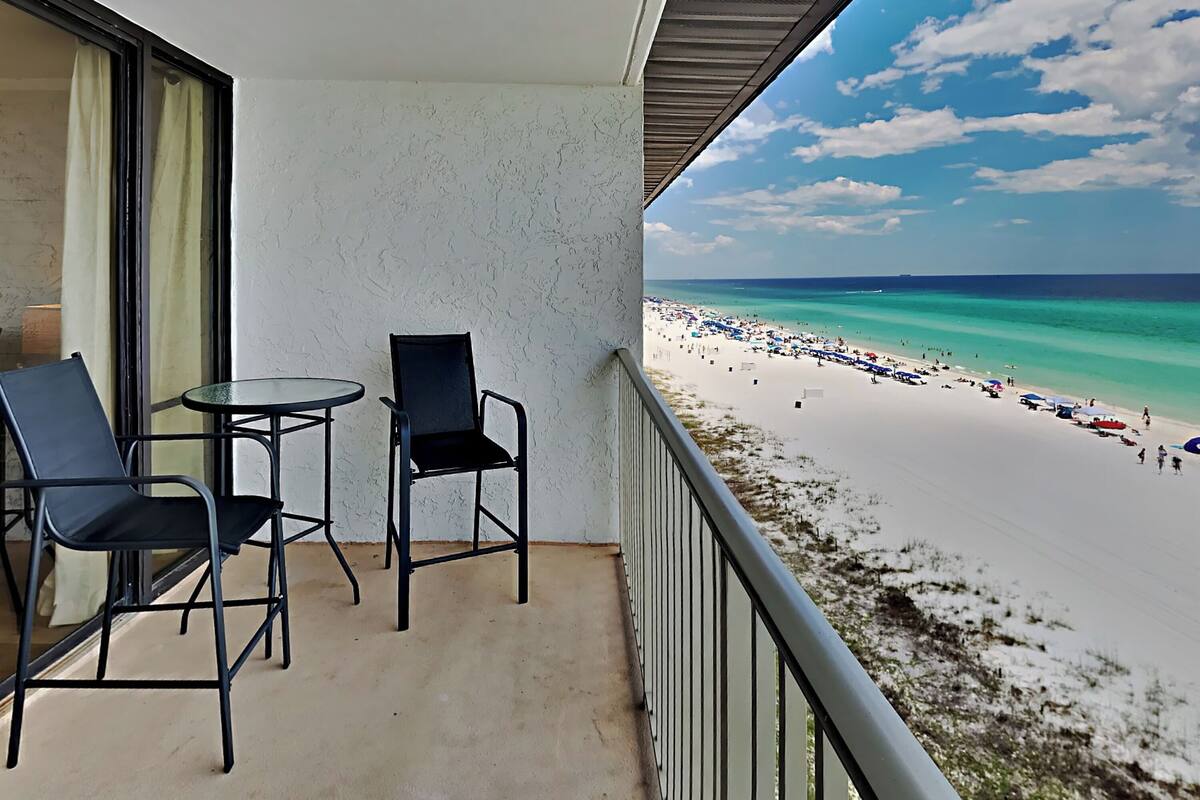 Ein Balkon mit zwei Stühlen und einem kleinen Tisch, mit Blick auf einen Strand mit Menschen und Sonnenschirmen.