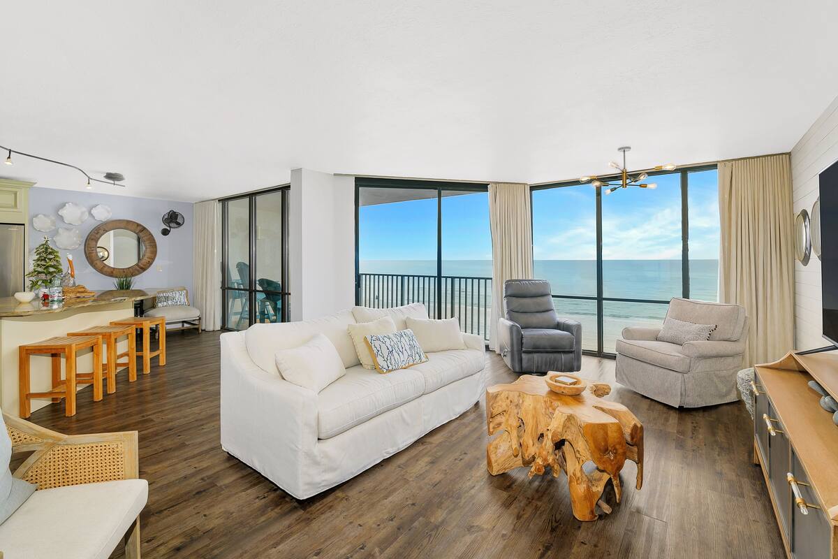 Ein geräumiger Wohnbereich mit einem weißen Sofa, zwei Sesseln, einem Holztisch und einem Essplatz. Große Fenster bieten Blick auf den Ozean.