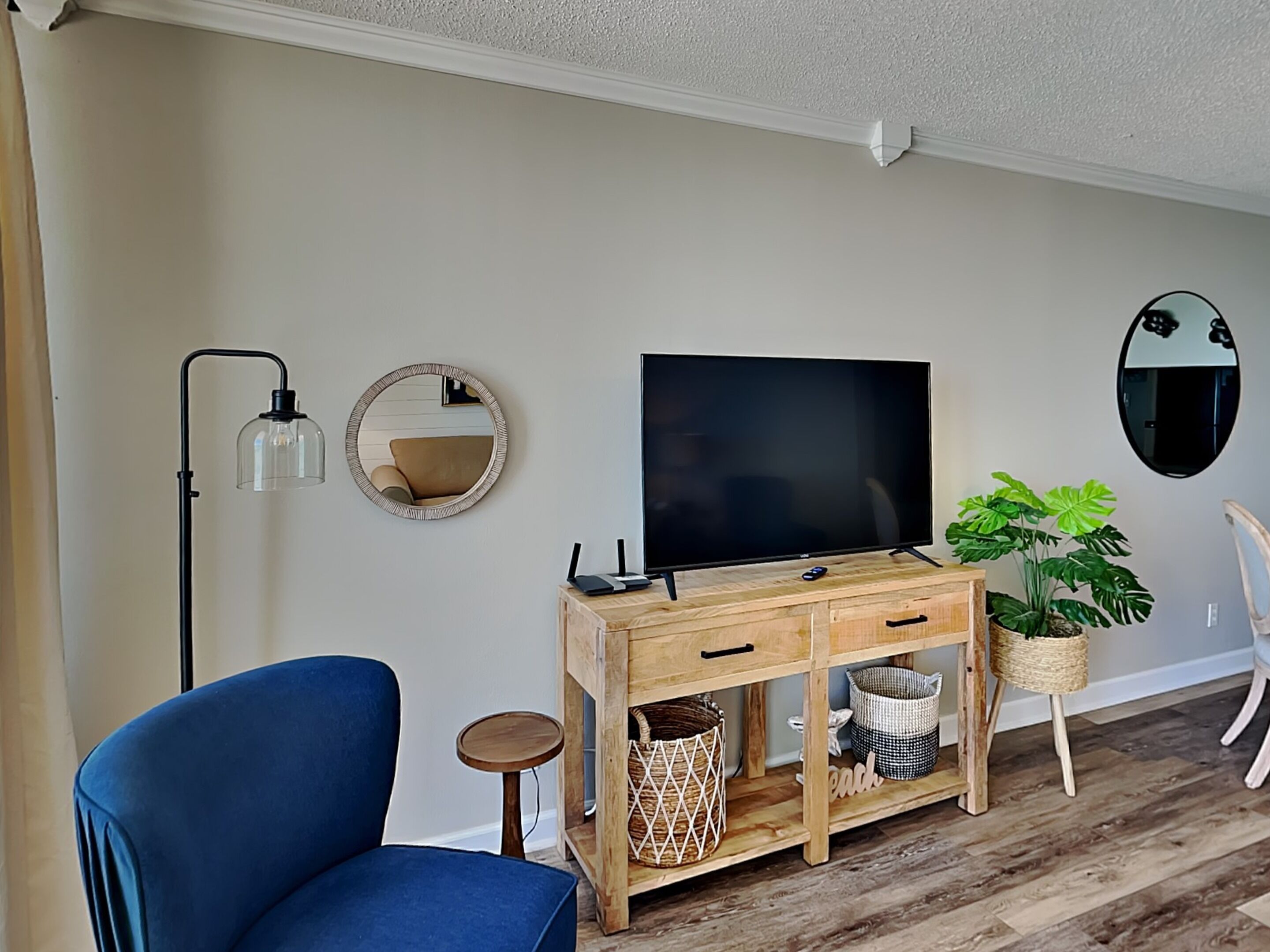 A living room with a TV on a wooden console, a blue armchair, a floor lamp, and two wall mirrors.