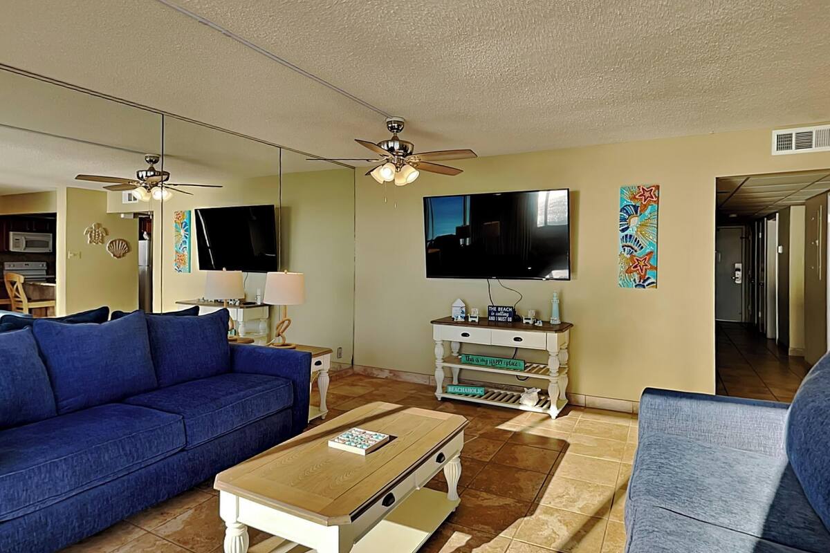 Ein Wohnzimmer mit einem blauen Sofa, einem Holztisch, einem Fernseher und einem Deckenventilator.