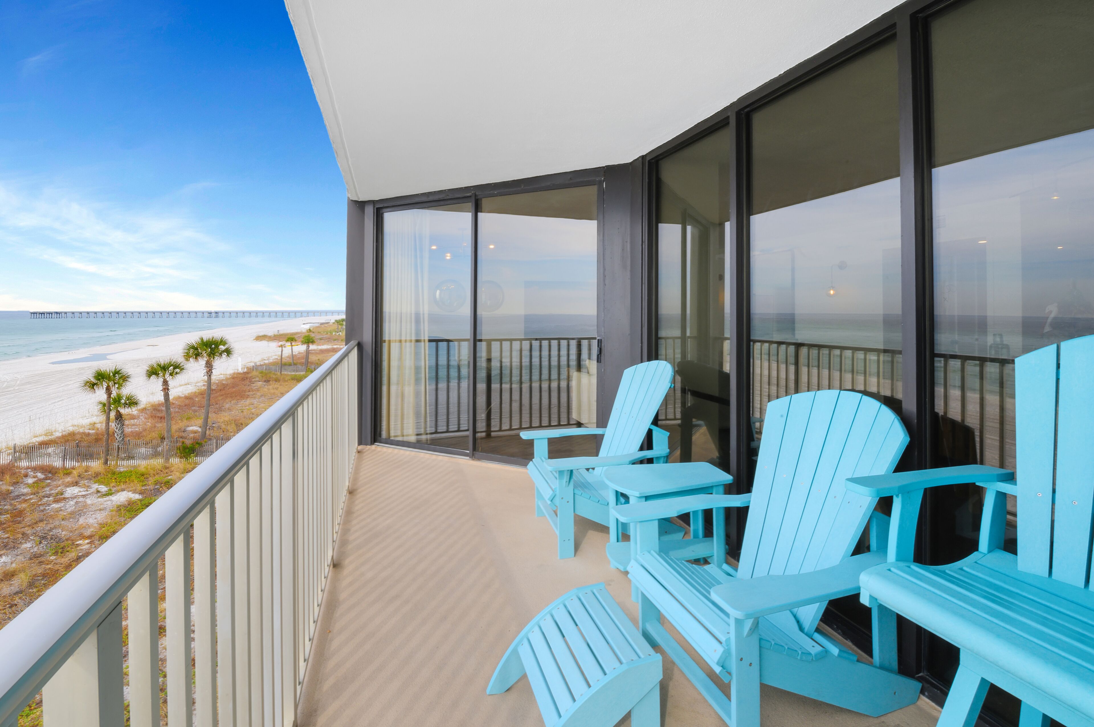A balcony with blue Adirondack chairs, a glass door leading to a beach view, and a wooden deck.