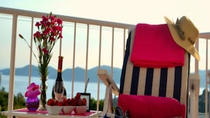 Un balcón con una silla, una mesa con una botella, un jarrón de flores y un cuenco de fruta.
