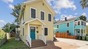 Une maison jaune à deux étages, avec une porte bleue et des bordures blanches, une allée pavée et une clôture en bois.