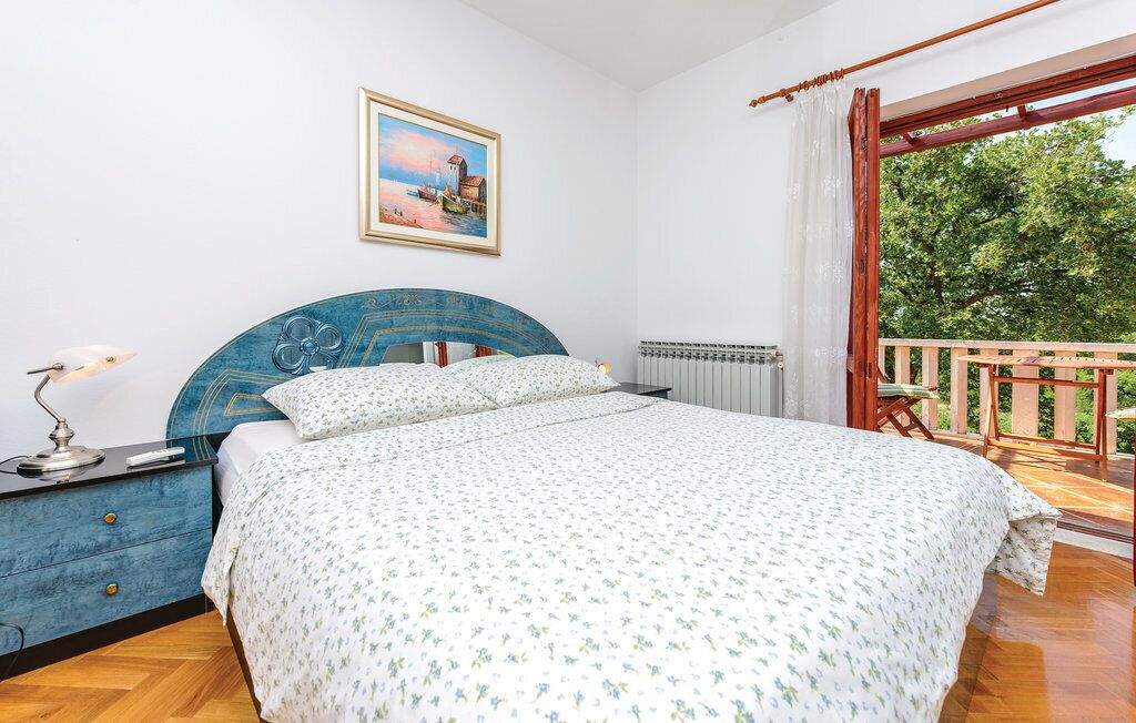 A bedroom with a bed, a nightstand, a lamp, a radiator, and a balcony with a view of trees.