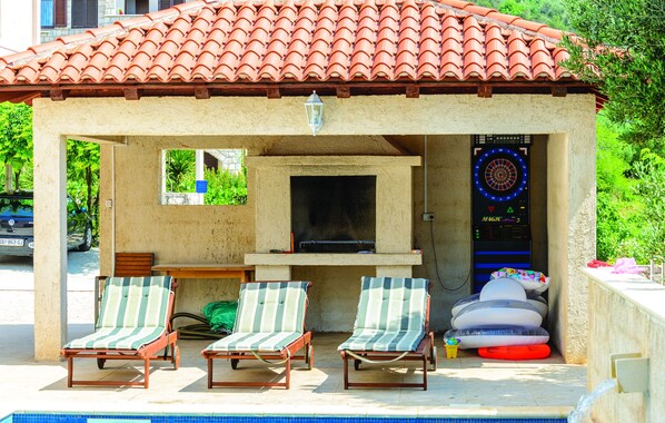 A covered outdoor area with a barbecue, two lounge chairs, and a poolside setting. - Villa Anka (Dubrovnik)