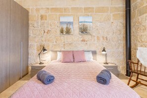 Une chambre avec un mur en pierre, un lit avec une literie rose, deux tables de chevet avec des lampes et une chaise.