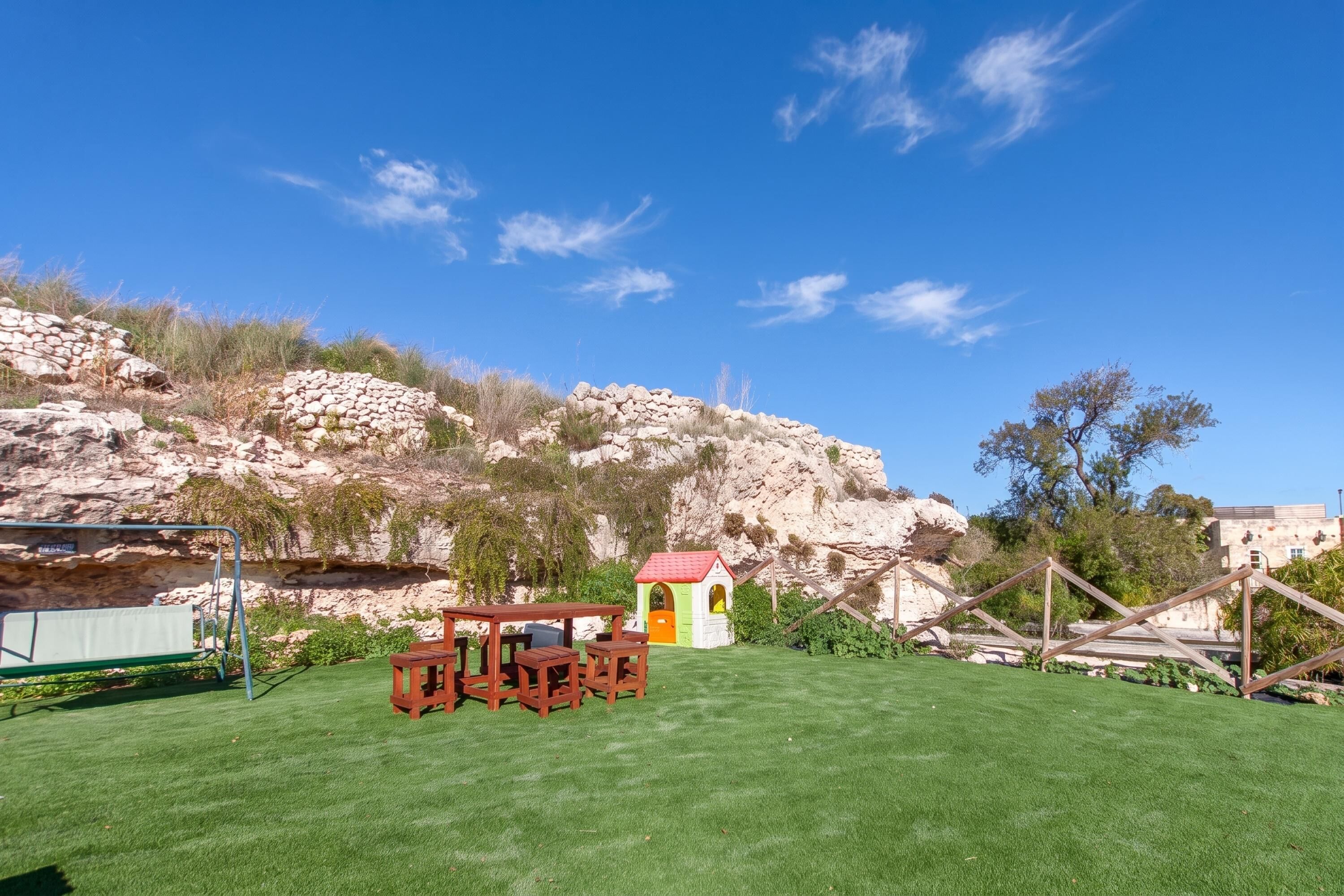 A grassy outdoor area with a small wooden table and chairs, a playhouse, and a rocky hillside in the background.