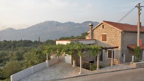 A stone house with a red-tiled roof, a small porch, and a view of mountains in the background. - Rilović House (Konavle)