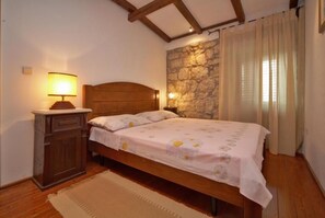 Un dormitorio con cama de madera, una mesita de noche con lámpara, pared de piedra y una ventana con cortinas.