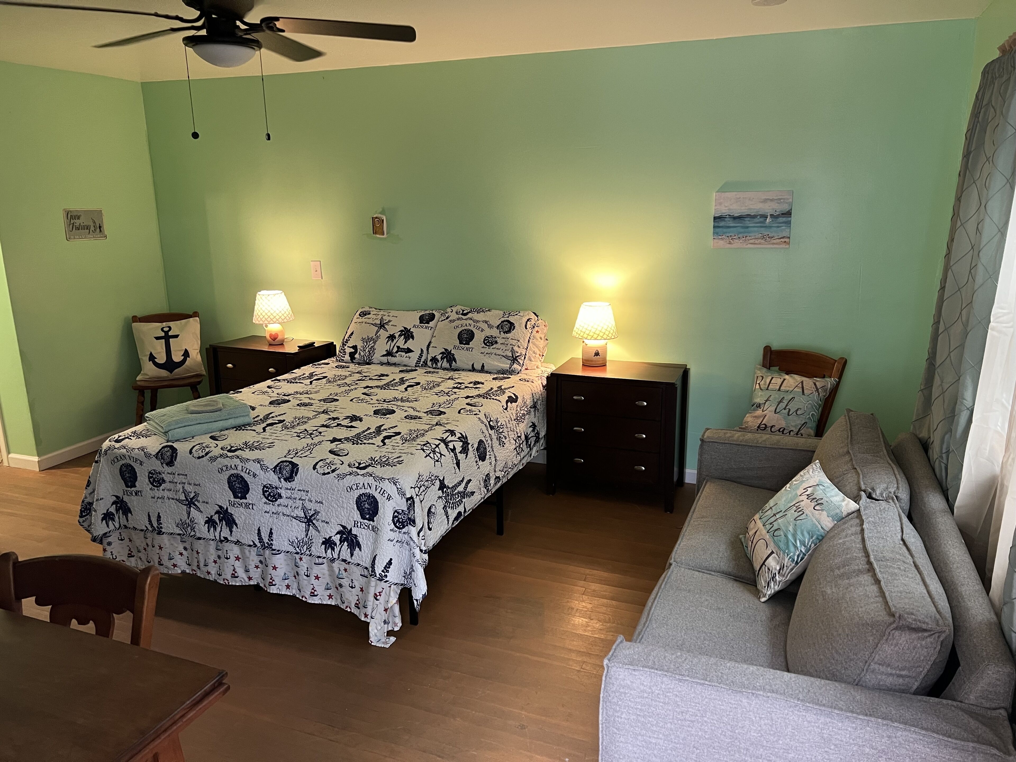 A bedroom with a bed, two nightstands, a ceiling fan, a gray sofa, and a wooden table.