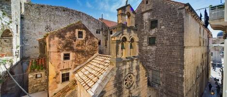 A narrow street with historic stone buildings, including a church with a bell tower and a small bell.