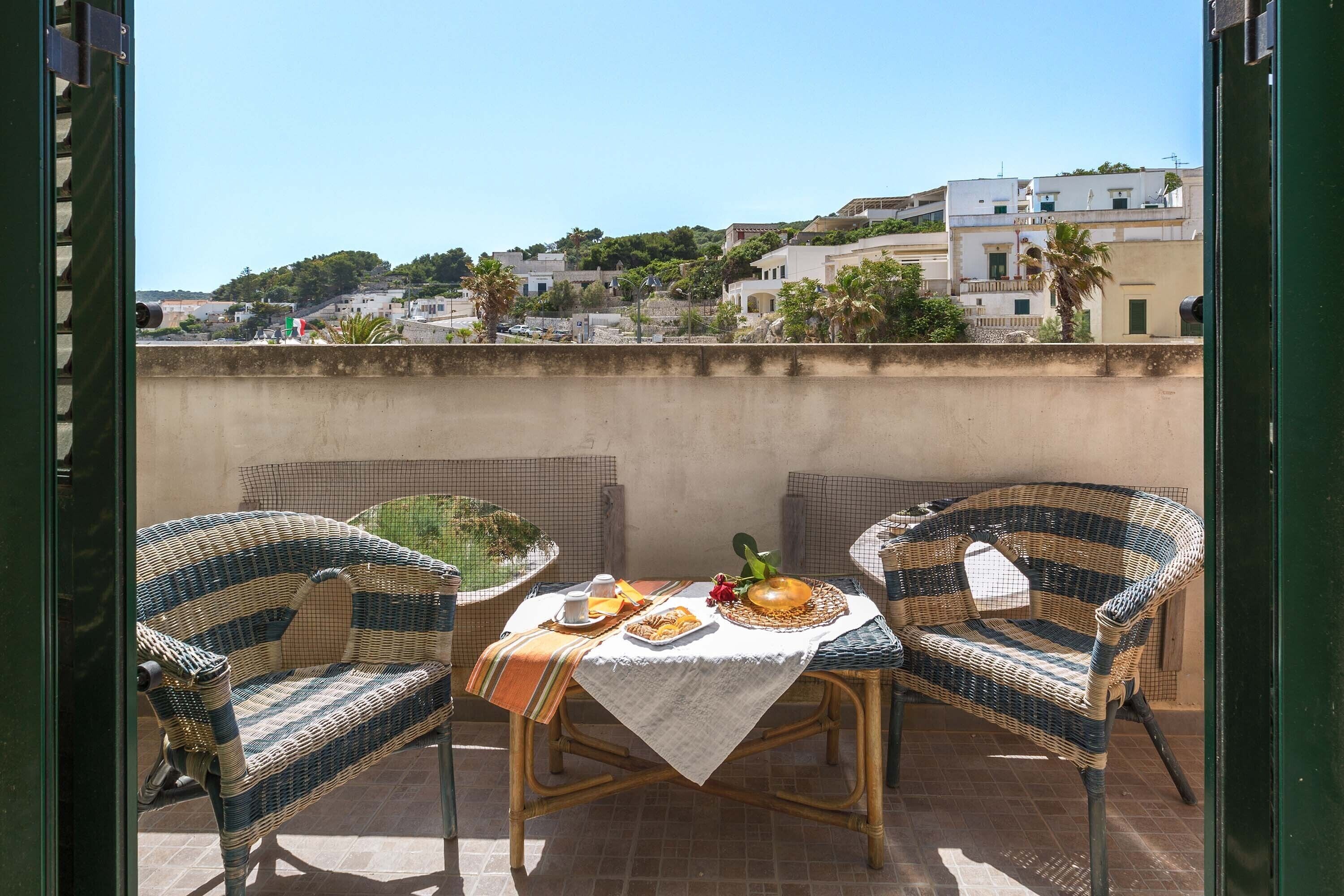 A balcony with wicker chairs, a table with a tablecloth, and a view of a hillside town.
