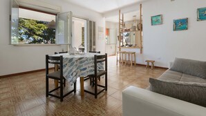 A dining area with a table set for four, a sofa, and a view of greenery outside. - 2718 Villetta Soffio di Mare by Barbarhouse (Melendugno)
