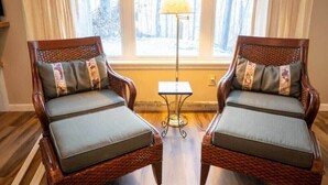Två fåtöljer av rotting med vadderade sitsar och ryggstöd, placerade framför ett fönster med utsikt över träd.