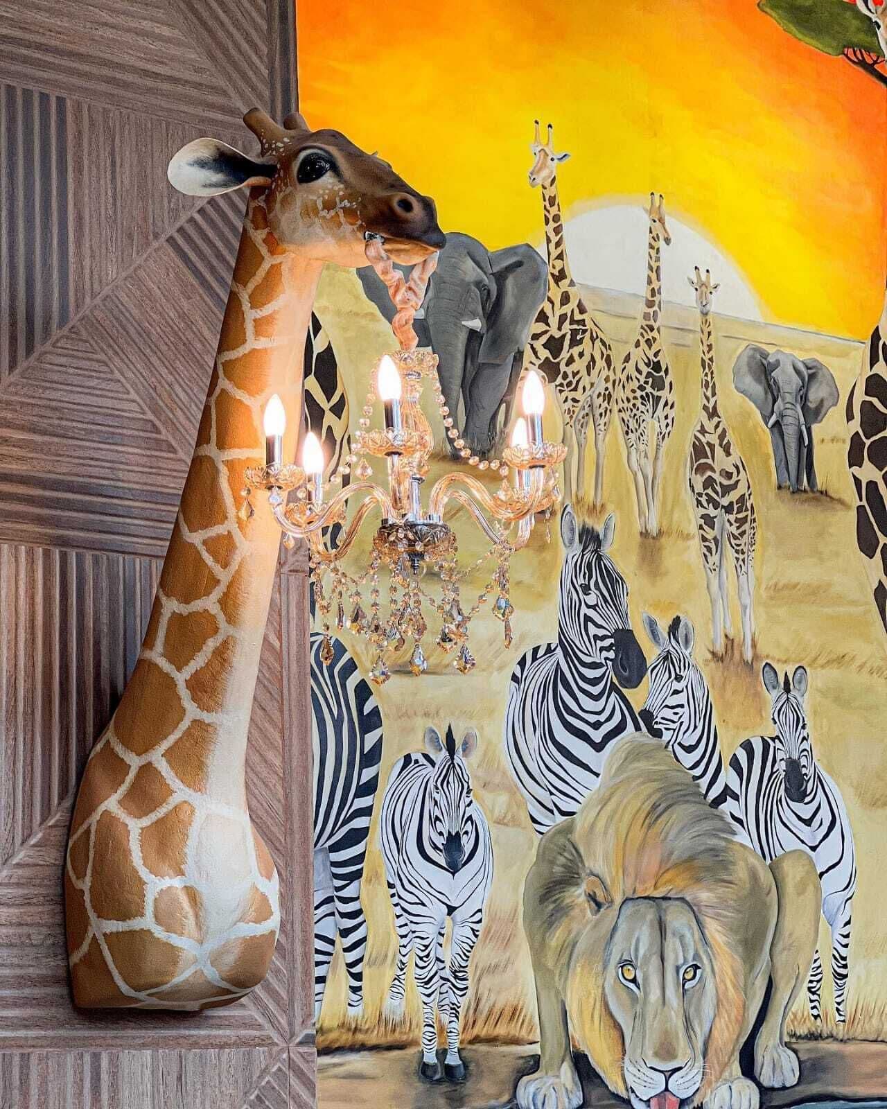 A giraffe sculpture with a chandelier, zebras, and a lion in a room with patterned walls.