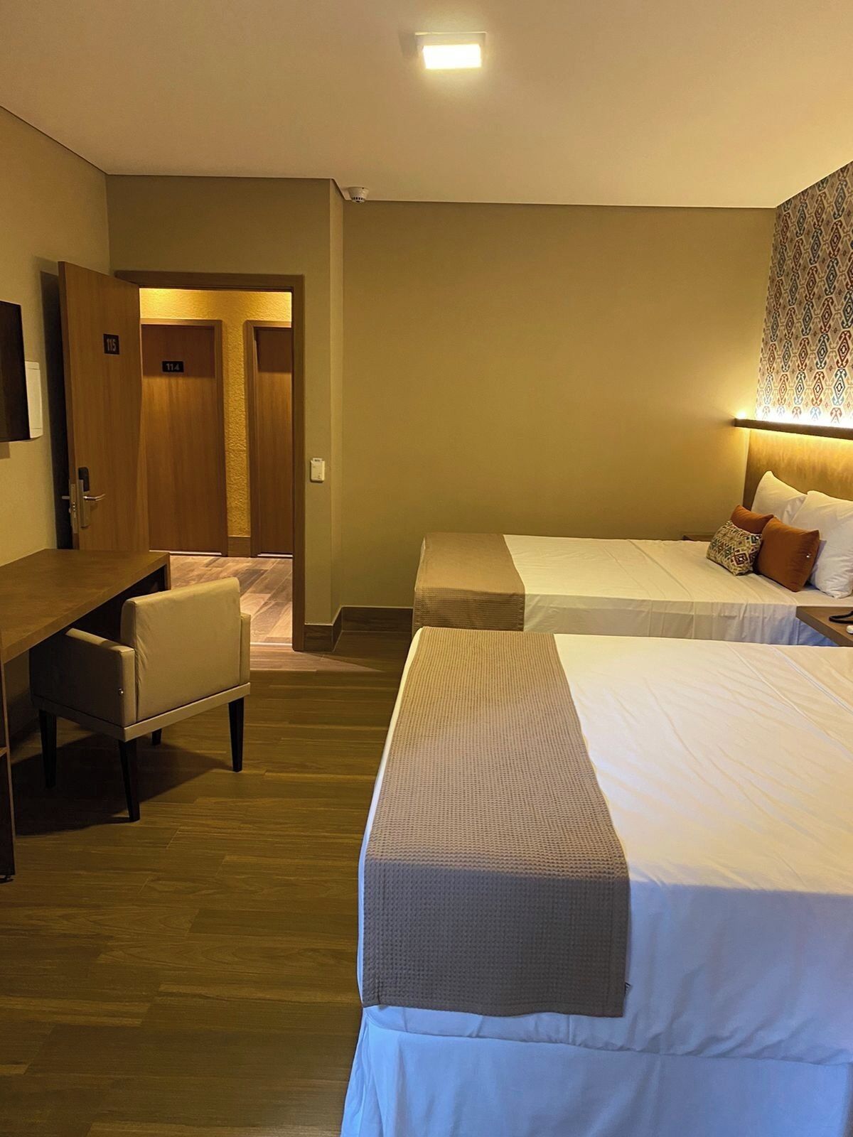 A hotel room with two beds, a desk, and a chair. There is a door leading to another room.
