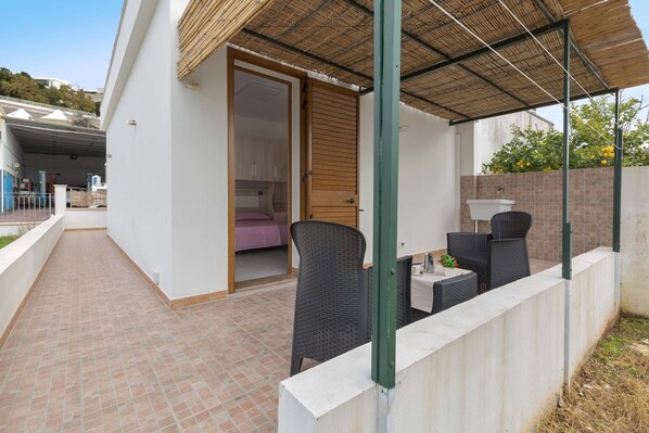 A covered outdoor seating area with wicker furniture, a concrete wall, and a door leading to a room with a bed. - 2215 Villetta Enea - Mono 2 by Barbarhouse (Castro)