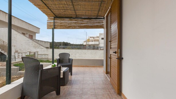 Ein überdachter Balkon im Freien mit Korbmöbeln, gefliestem Boden und Blick auf Gebäude im Hintergrund.