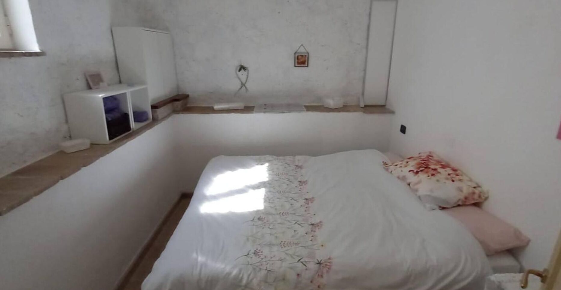 Una habitación pequeña y sencilla con una cama, un mueble blanco pequeño y una estantería en la pared.