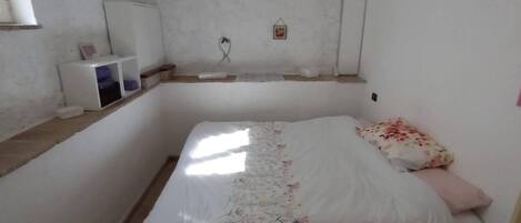 Una habitación pequeña y sencilla con una cama, un mueble blanco pequeño y una estantería en la pared.