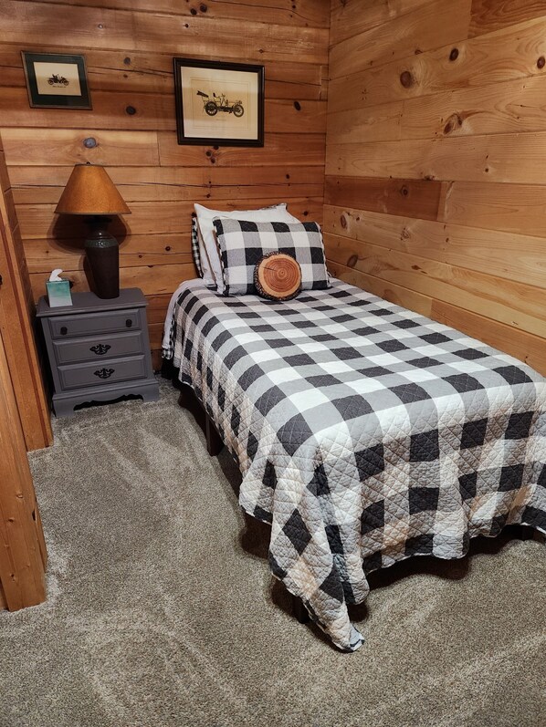 A cozy bedroom with a checkered bedspread, a nightstand with a lamp, and framed pictures on the wall. - Travelers Rest Cabin (Warm Springs)