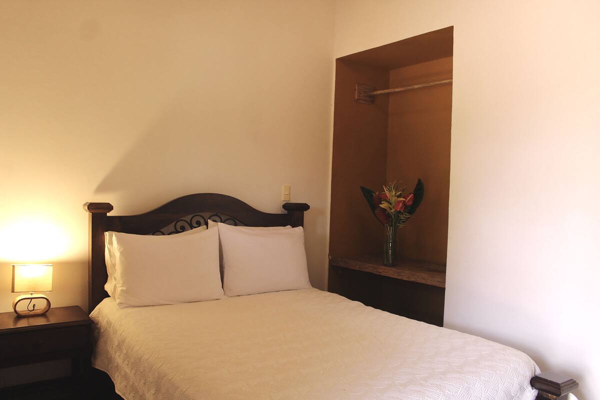 A neatly made bed with a wooden headboard, a bedside lamp, and a small shelf with a vase of flowers.