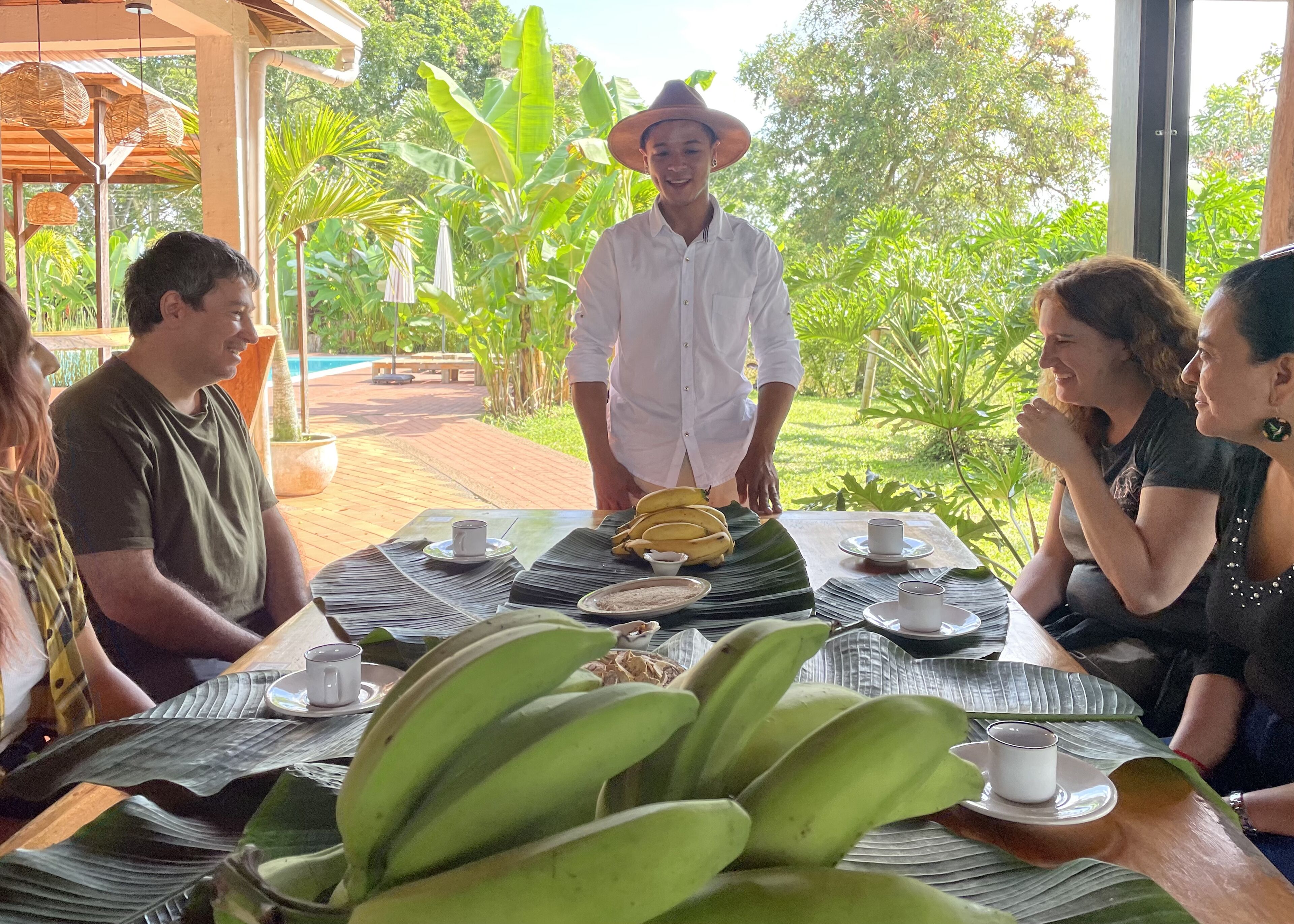Un groupe de personnes est assis à une table à l’extérieur, avec un bananier au premier plan, une terrasse en bois et une végétation luxuriante en arrière-plan.