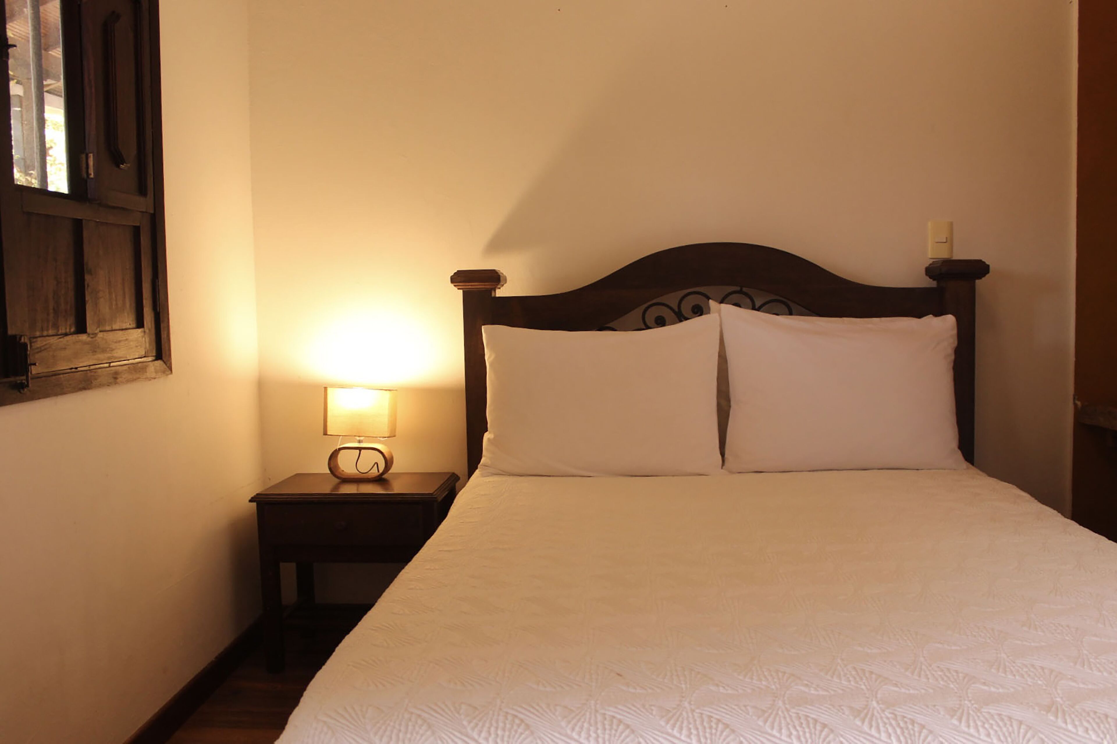 Una cama bien hecha con sábanas blancas y un cabecero de madera, una mesita de noche con una lámpara, y una ventana con marco de madera.