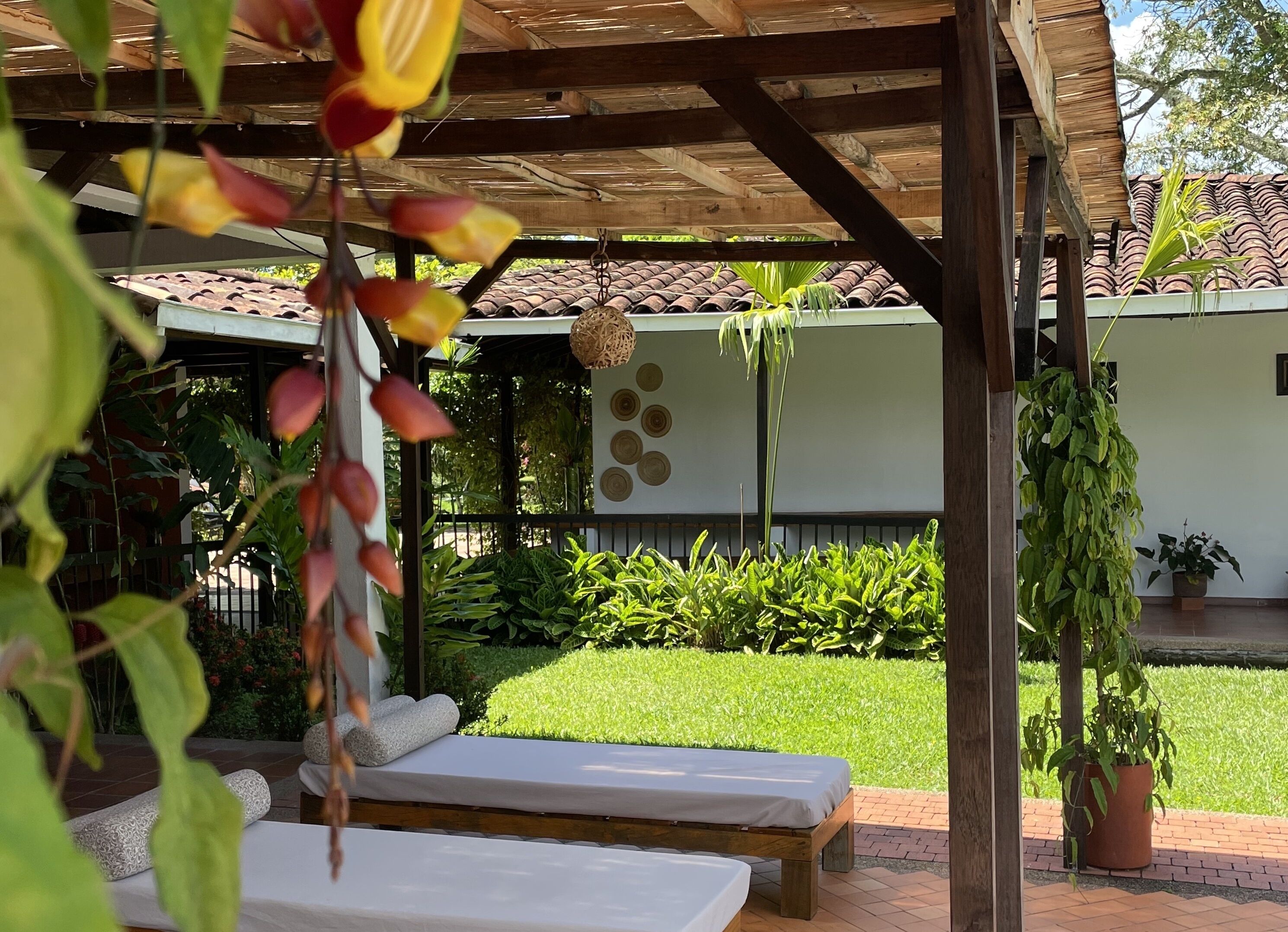 A covered outdoor seating area with two lounge chairs, a wooden ceiling, and a garden with various plants and a tiled roof in the background.