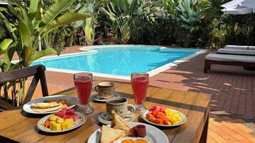 Una mesa de desayuno con platos de comida, un vaso de jugo y vista a una piscina.