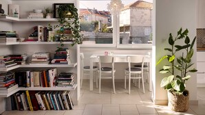 Una habitación bien organizada con estanterías blancas llenas de libros, una mesa de comedor con sillas y un amplio ventanal con vistas a edificios.