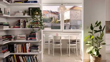 A well-organized room with white shelves filled with books, a dining table with chairs, and a large window offering a view of buildings outside.