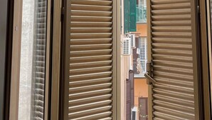 A view through a window with closed blinds, revealing a narrow street with buildings and balconies. - Cavalieri Affittacamere (Rome)