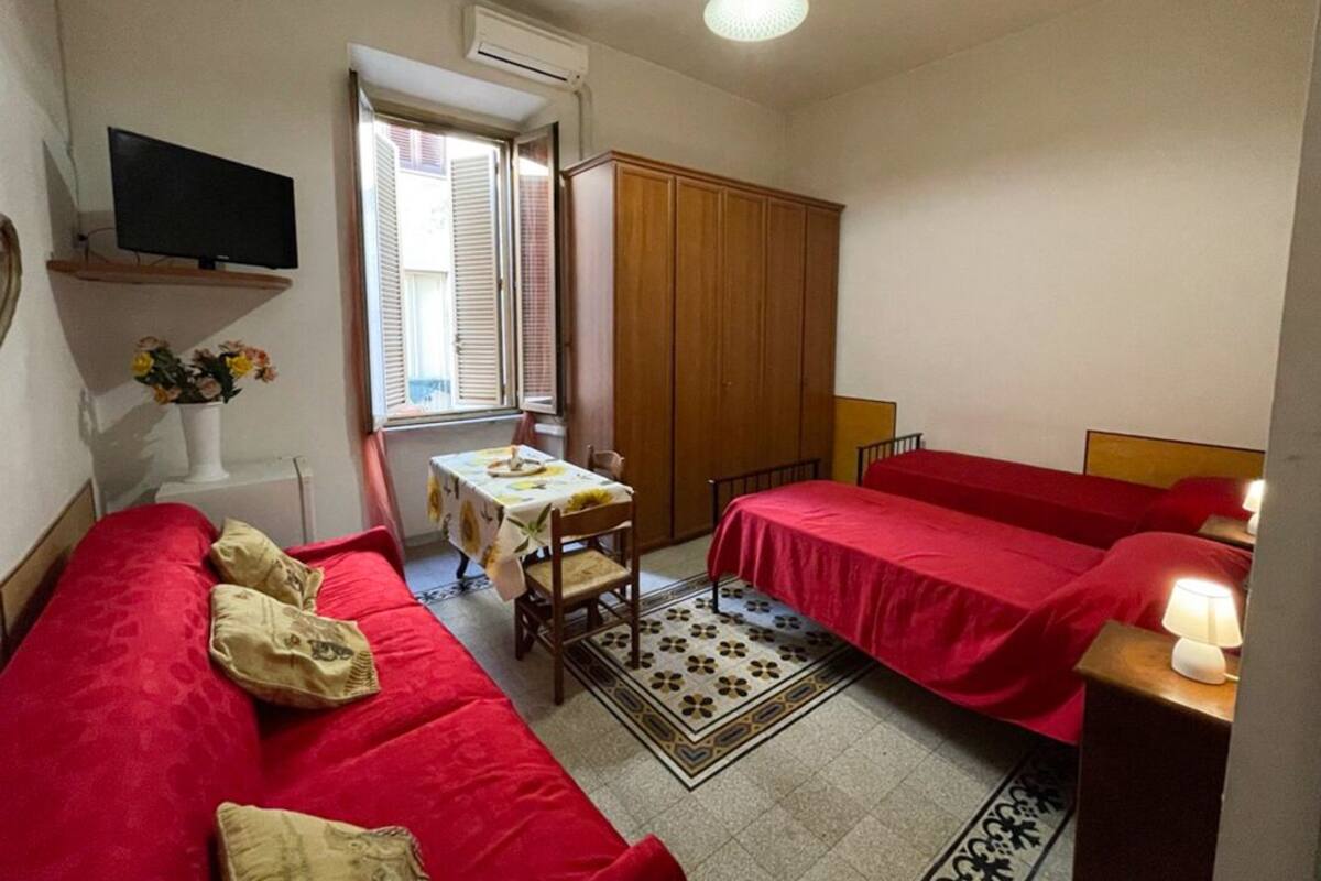 A small, cozy room with a red sofa, a bed with a red bedspread, a small table with a floral tablecloth, and a television mounted on the wall.