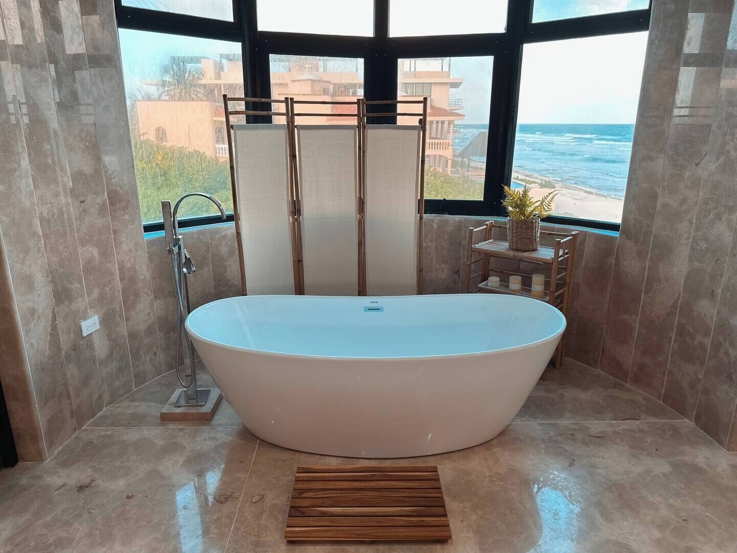 A modern bathroom with a freestanding bathtub, a wooden bench, and a view of the ocean through large windows.