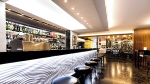 A modern bar with a long counter, black stools, and a variety of bottles on shelves.