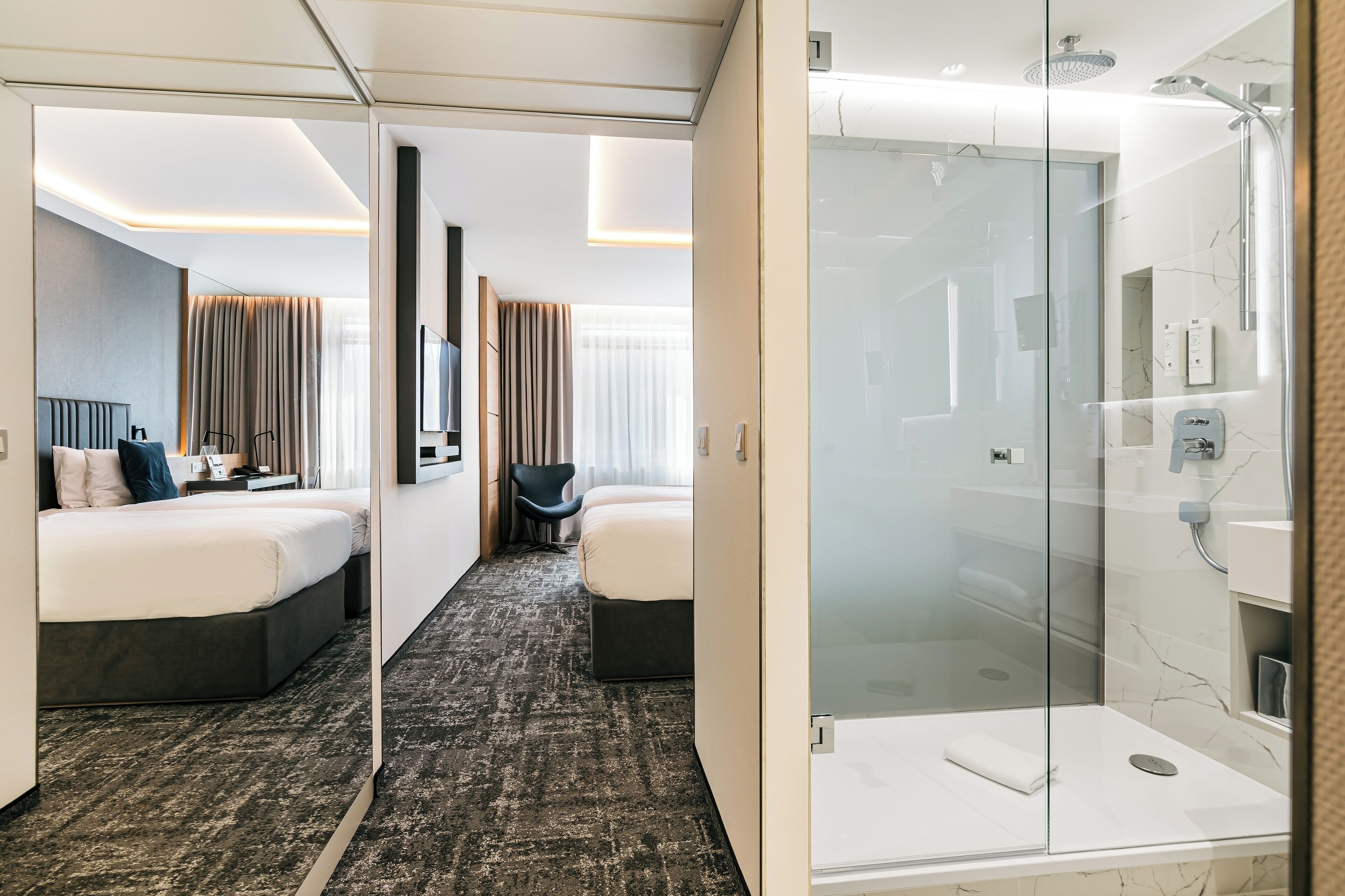 Habitación de hotel con dos camas, ducha y baño con paredes de vidrio.