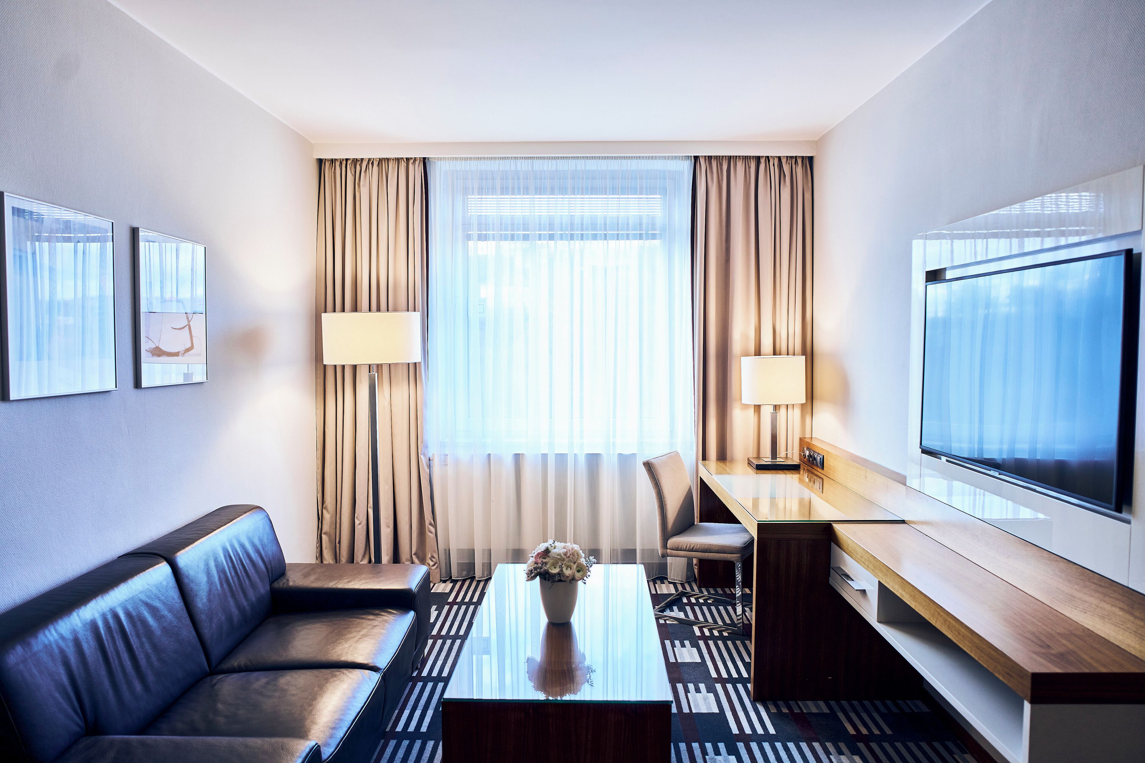 A modern hotel room with a sofa, a coffee table with a vase, a desk with a chair, a TV, and large windows with curtains.