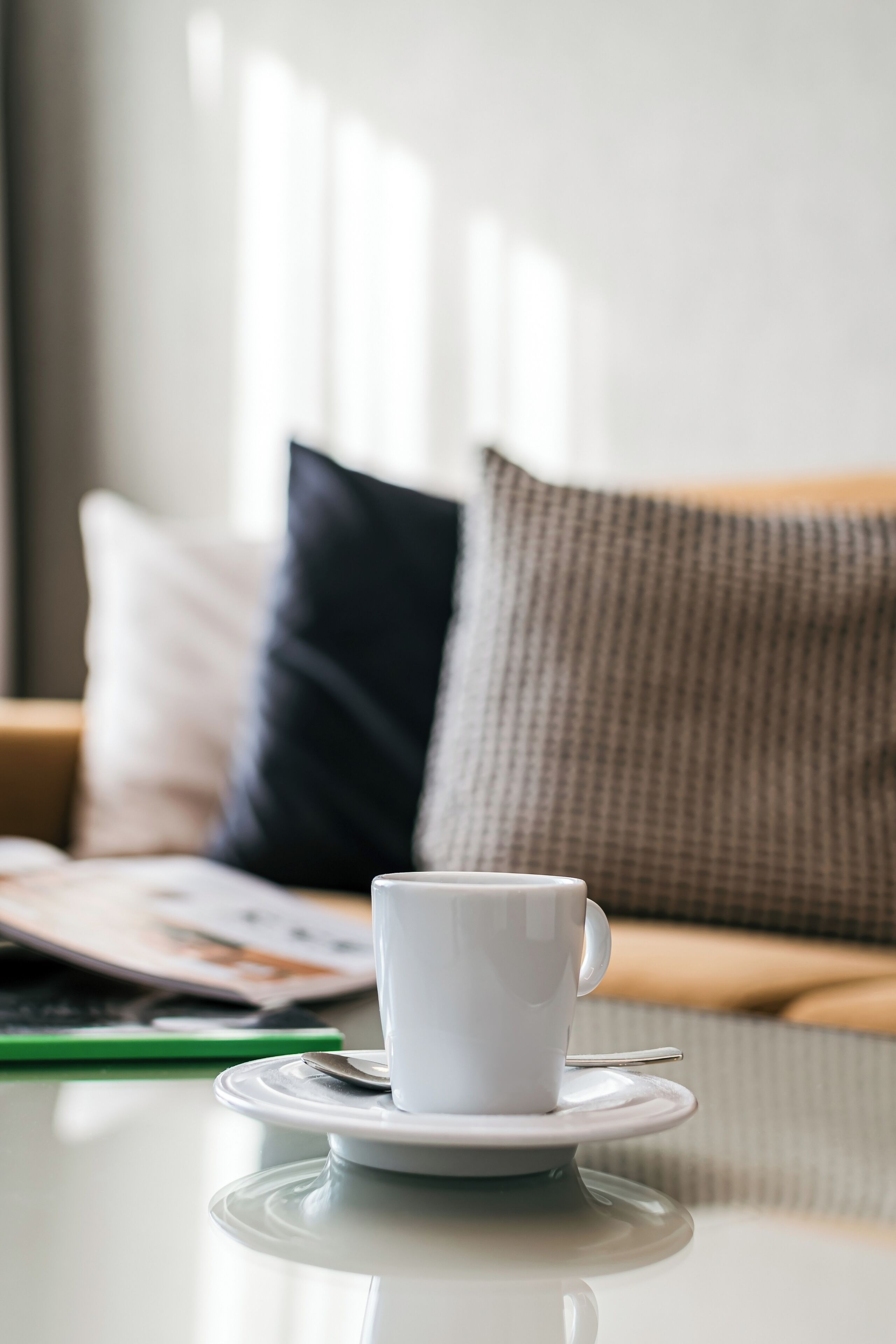 A white cup on a saucer on a table with a magazine and a pen beside it.