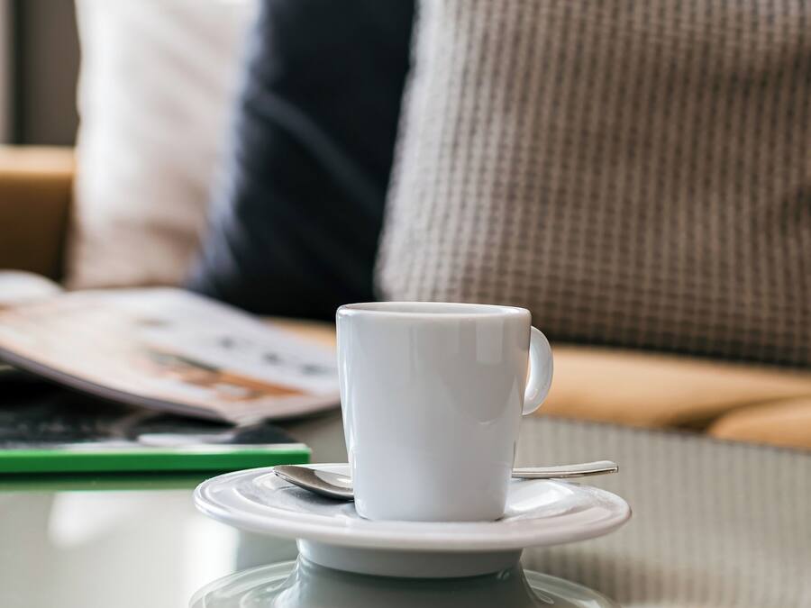 A white cup on a saucer on a table with a magazine and a pen beside it.