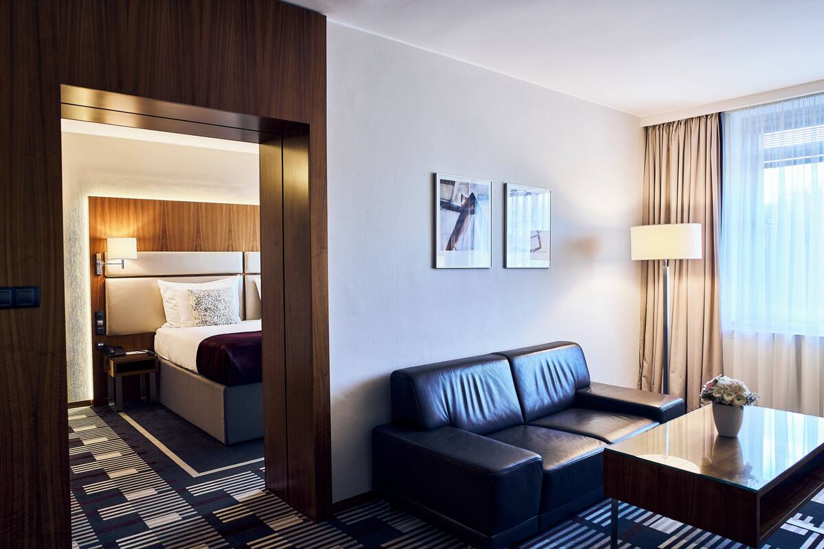 Hotellihuone, jossa on sänky, sohva, maljakkoon asetettu kahvipöytä ja kaksi kehystettyä kuvaa seinällä.