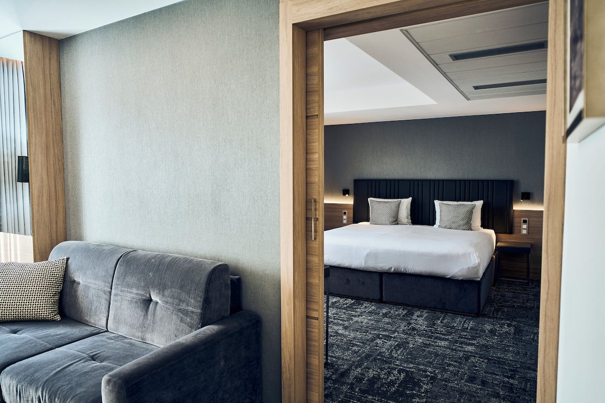 Moderni hotellihuone, jossa on harmaa sohva, siististi pedattu sänky tummalla päätyosalla ja puukehystetty oviaukko, joka johtaa toiseen huoneeseen.