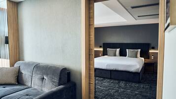 Una habitación de hotel moderna con un sofá gris, una cama bien hecha con cabecera oscura y un marco de puerta de madera que conduce a otra habitación.