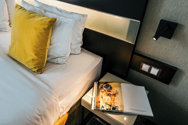 A neatly made bed with white and yellow pillows, a bedside table with a magazine, and a wall-mounted light fixture. - Hotel International Brno (Brno)