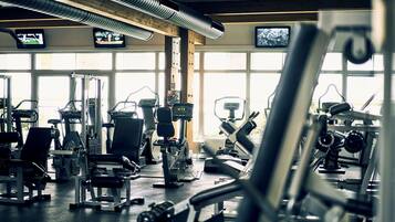 A gym with various exercise machines, large windows, and multiple TV screens on the wall.
